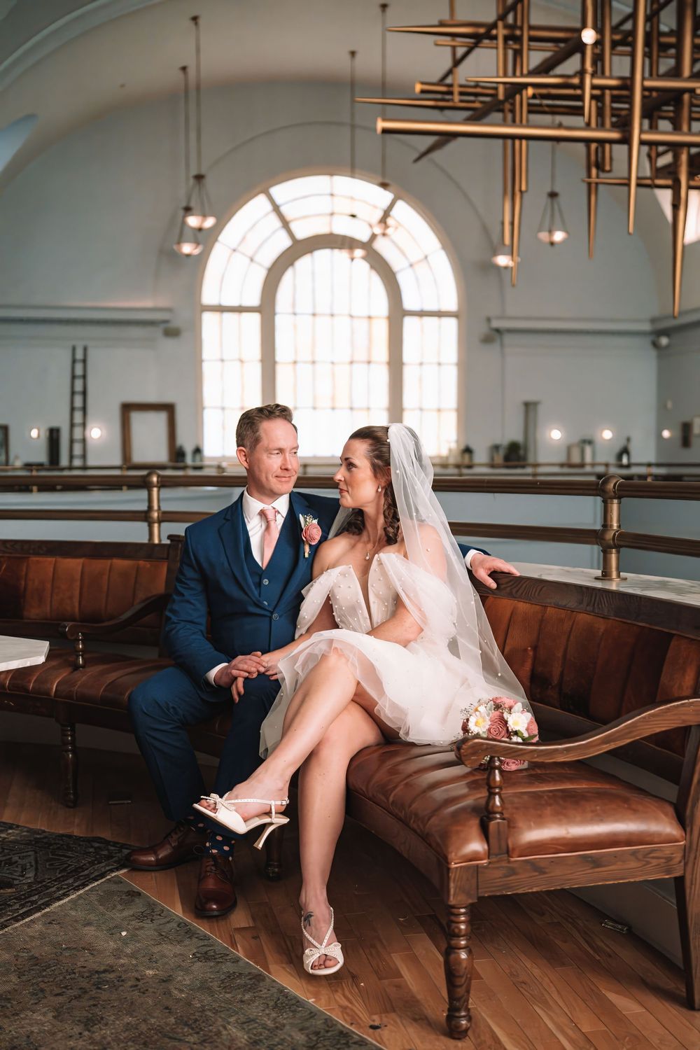 Couple sharing intimate moments on vintage leather couch in historic building with arched windows.