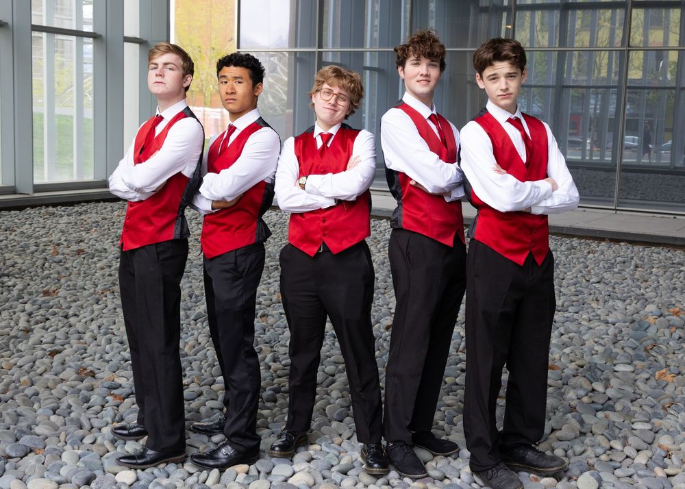 Group of young performers in formal attire sitting in a row outdoors.