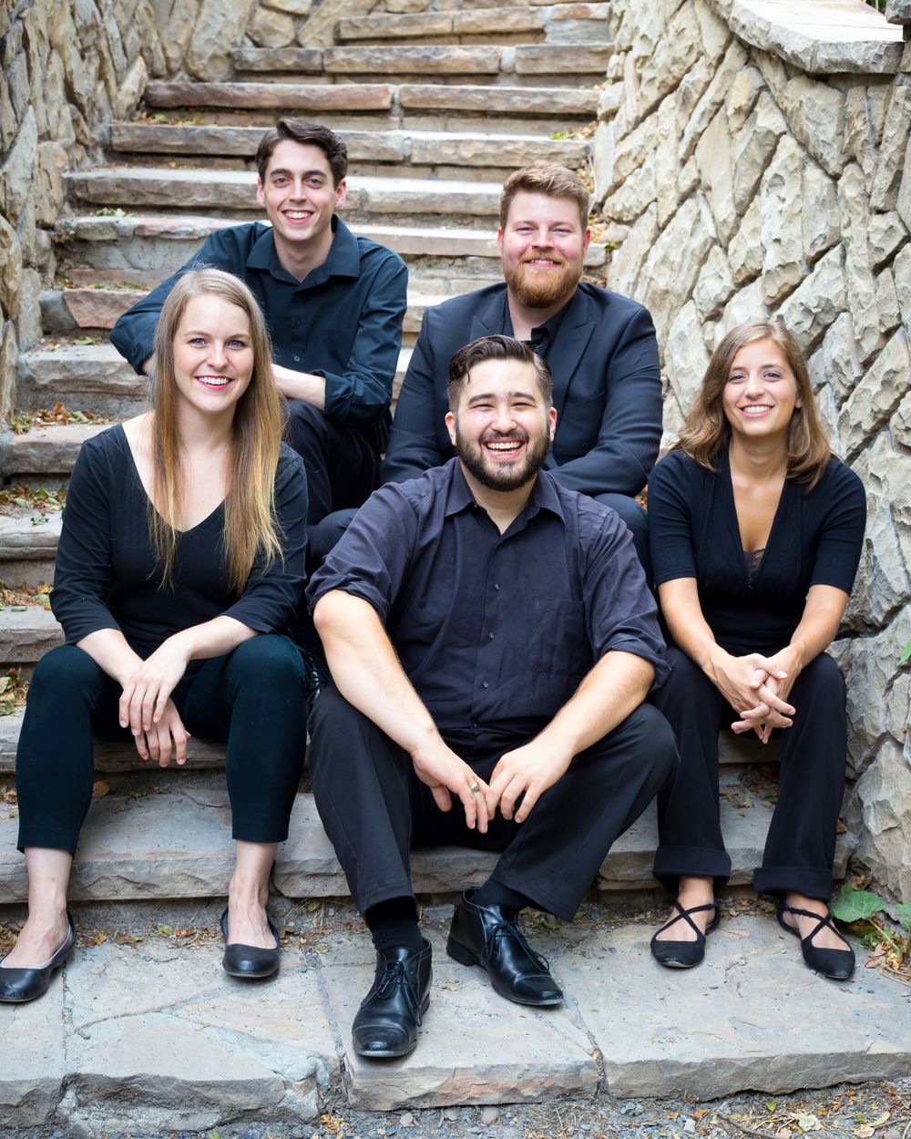 Group of performers in black attire sitting casually on stone steps outdoors.