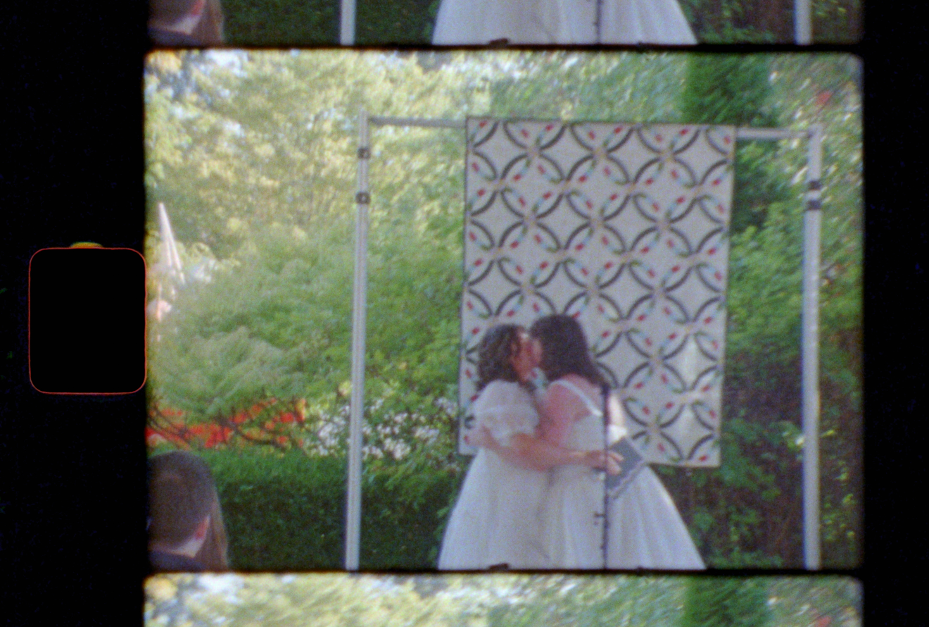 Two people in white dresses embrace during an outdoor wedding ceremony with a decorative backdrop panel.