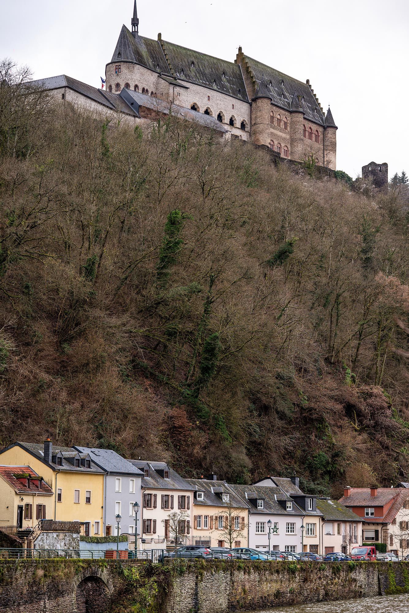 Historic Vianden Castle perches dramatically on a forested hillside above colorful houses along the riverside in Luxembourg.