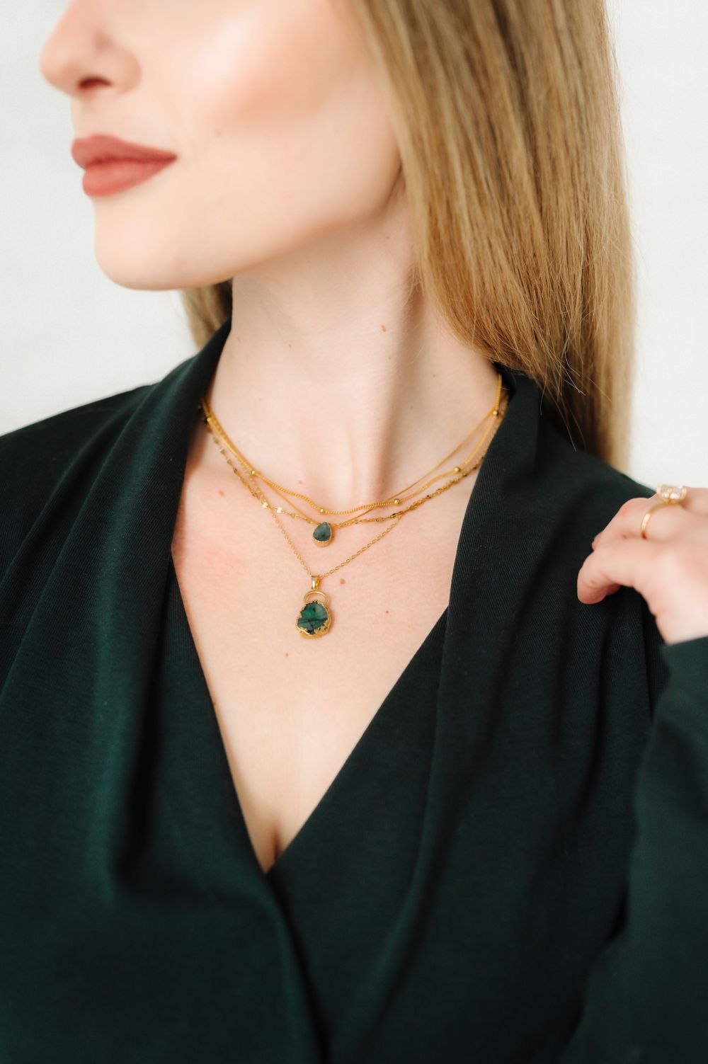Close-up of delicate gold necklaces with green gemstone pendants against dark green blouse.