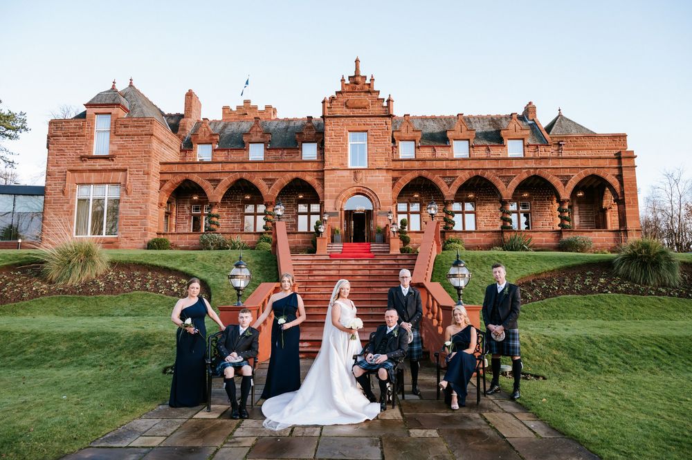 Wedding party poses in front of a historic red brick mansion on a grassy hillside during an outdoor celebration.