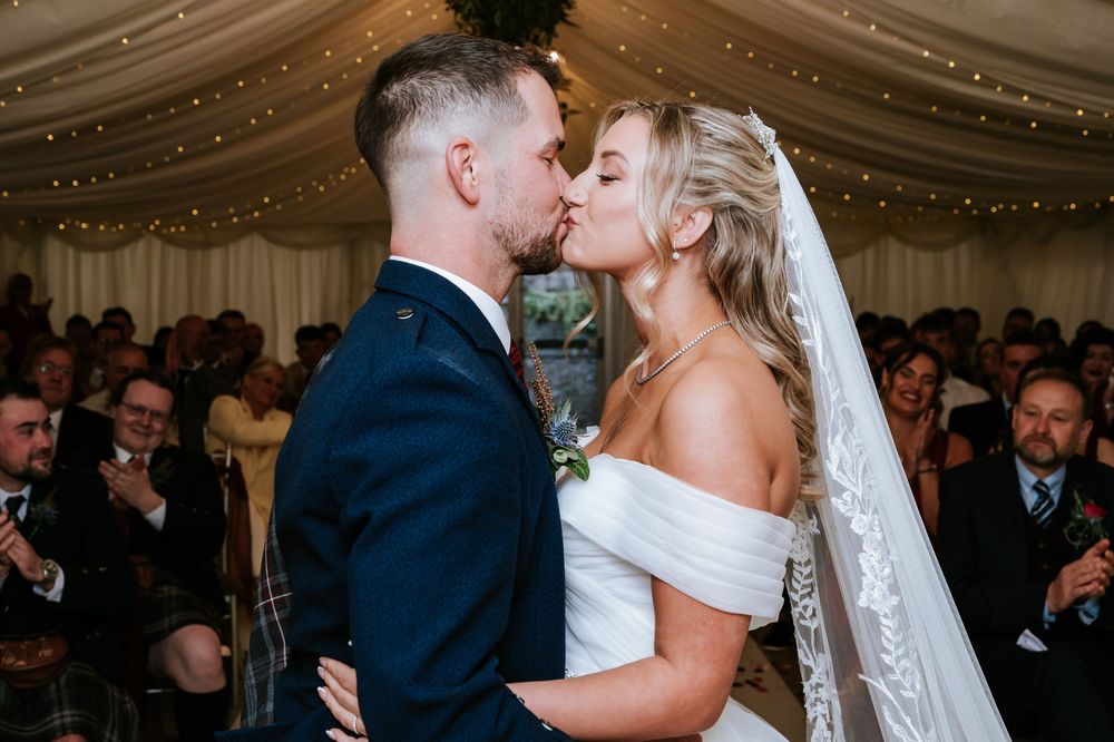 A romantic wedding ceremony moment as a couple shares their first kiss under a tent with twinkling lights and guests watching.