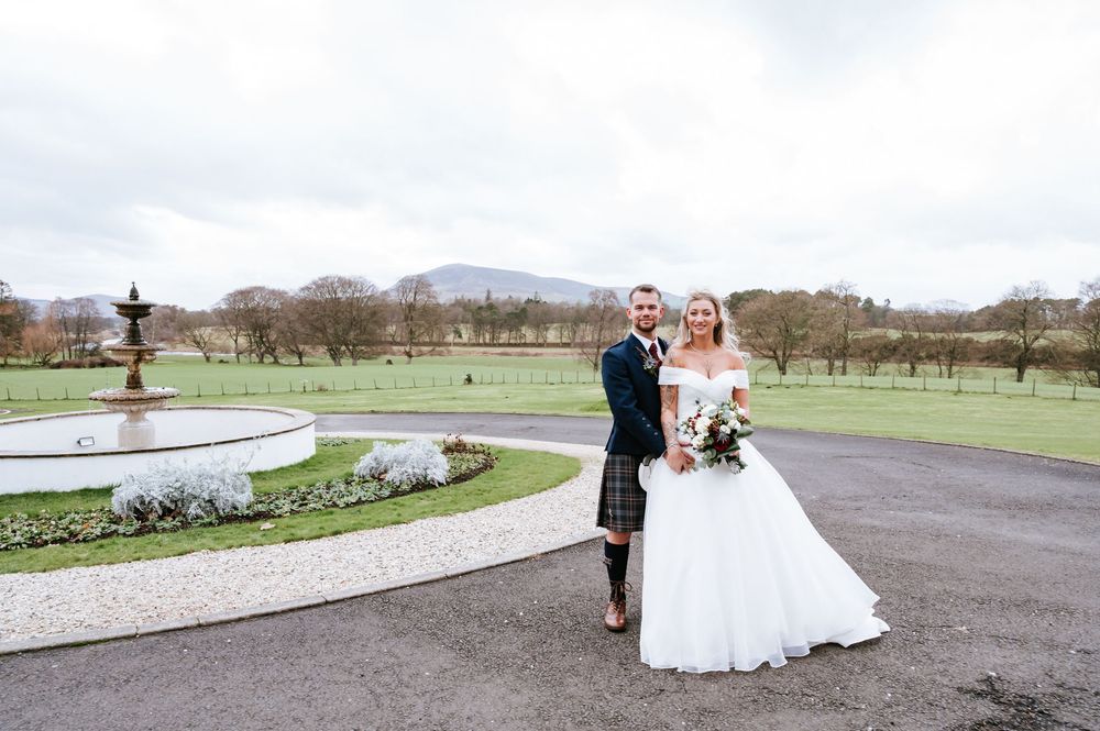 Couple in wedding attire poses along a curved driveway with mountain landscapes in the background.