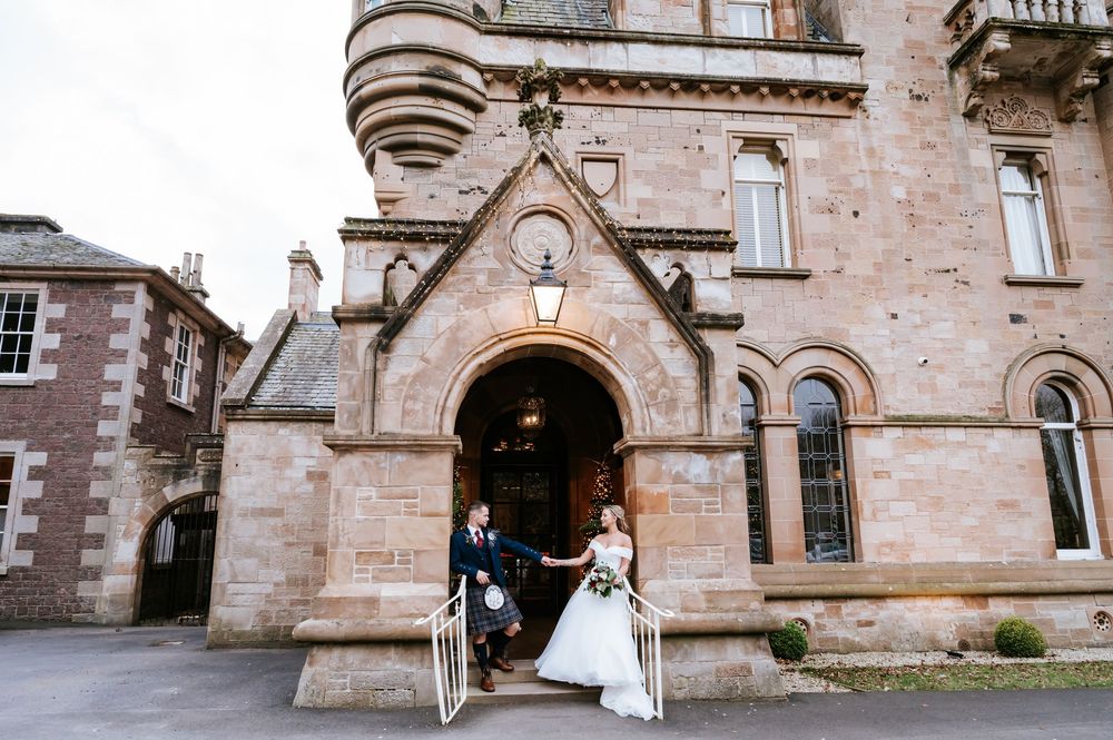 Wedding couple walks together through stone archway of historic building during outdoor ceremony.