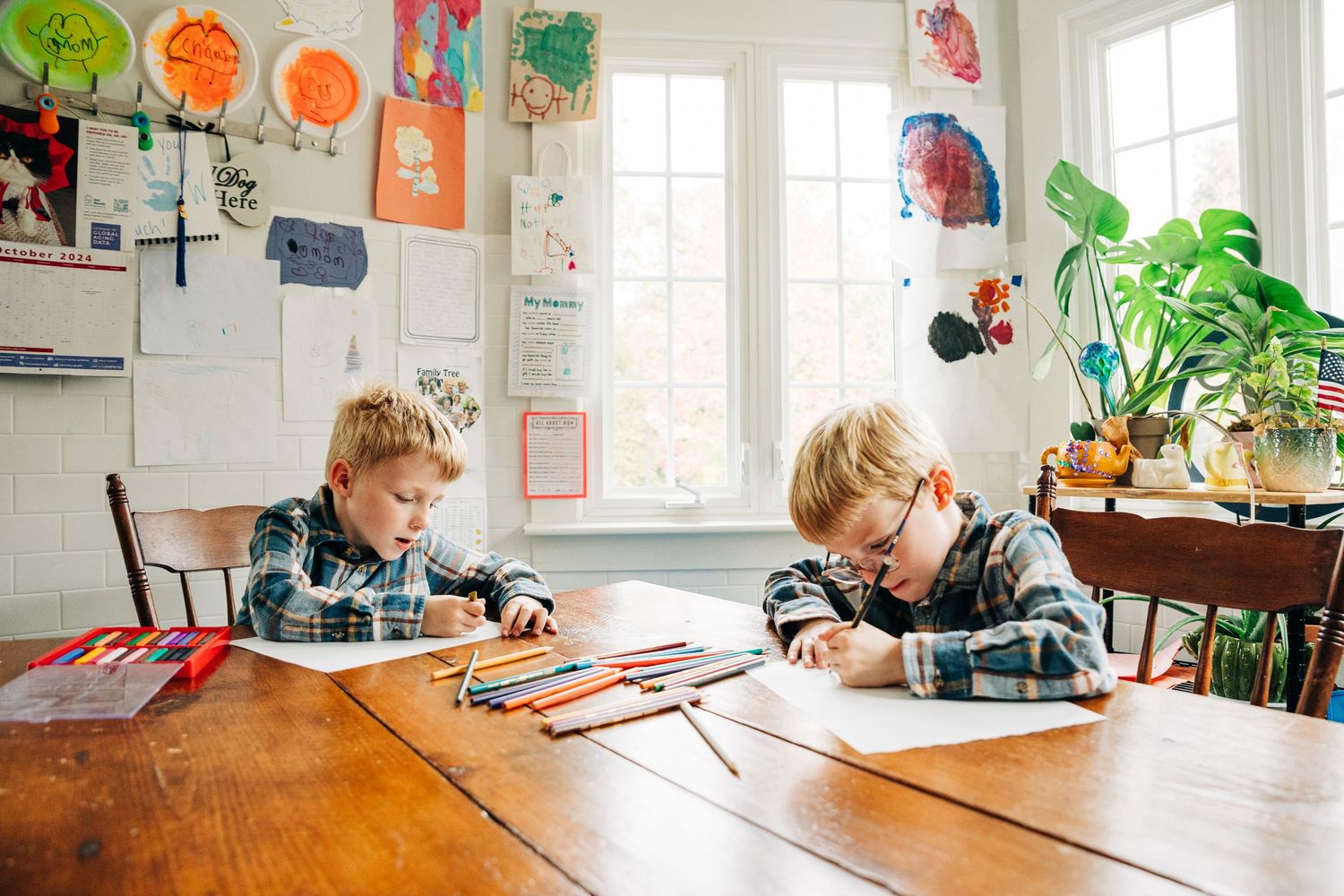 Twins work independently at a wooden table in a bright kitchen with colorful artwork and plants on display.