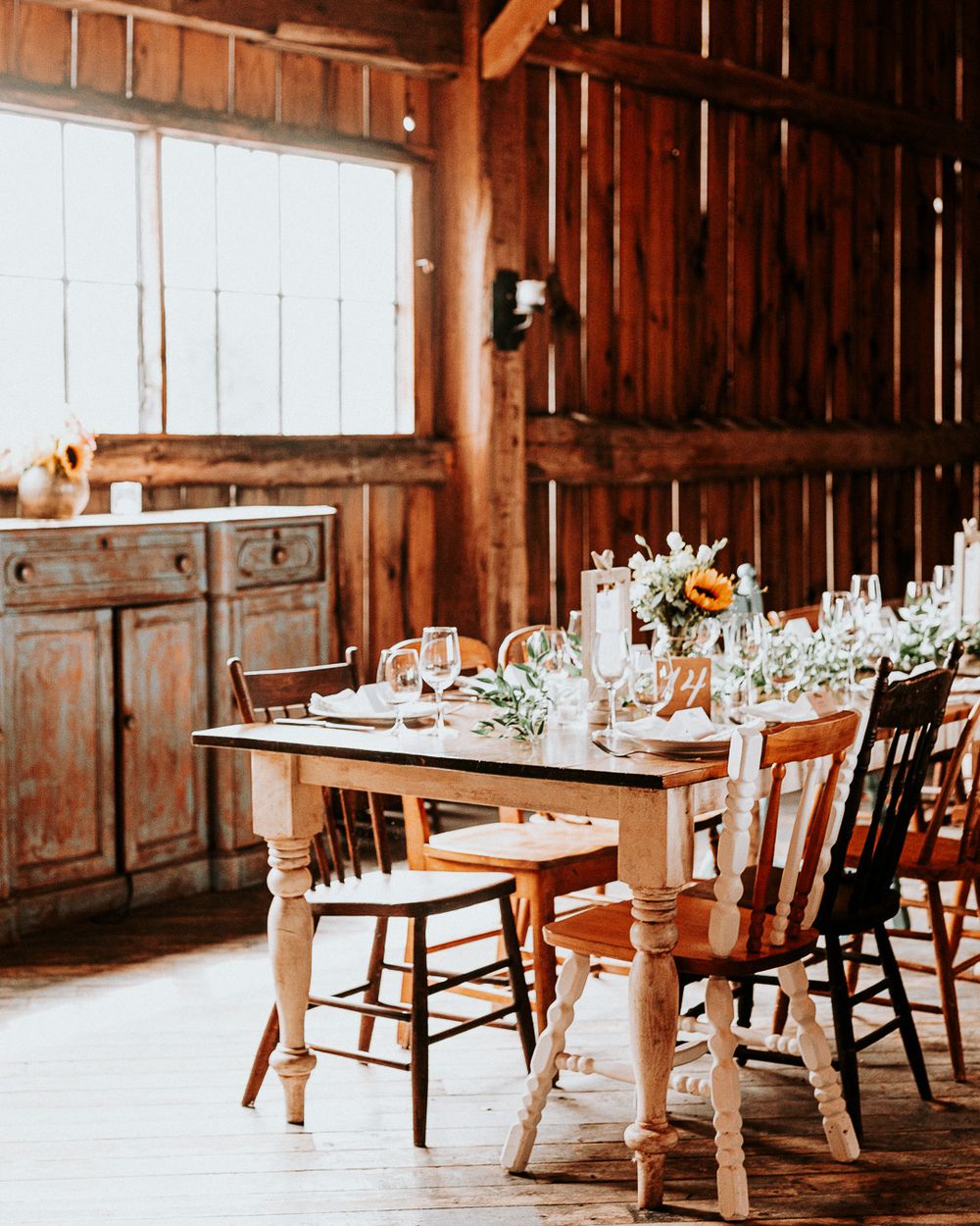 Rustic barn wedding reception setup with wooden table, chairs and elegant place settings in warm natural light.