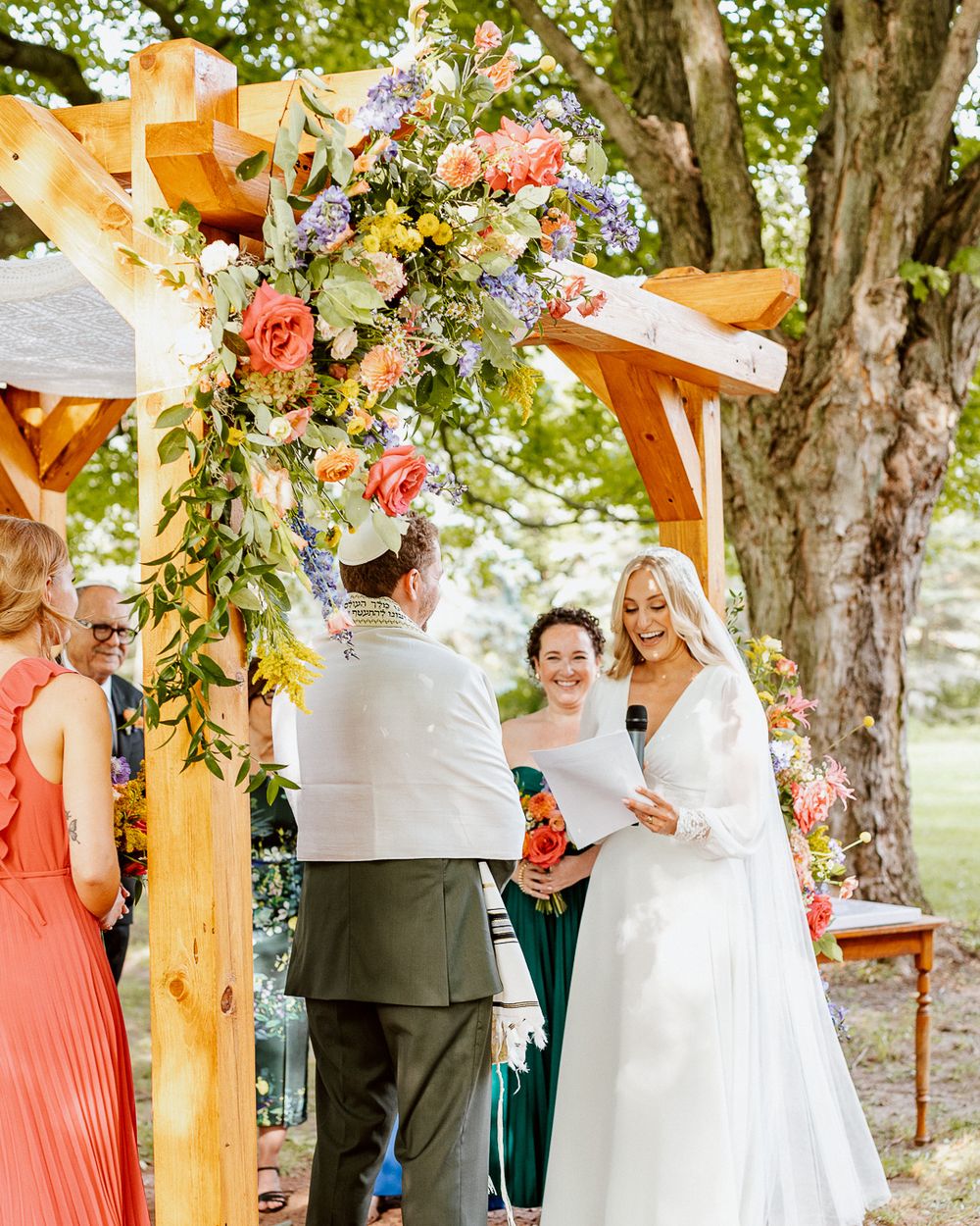 Outdoor wedding ceremony under wooden arch adorned with colorful floral arrangements.