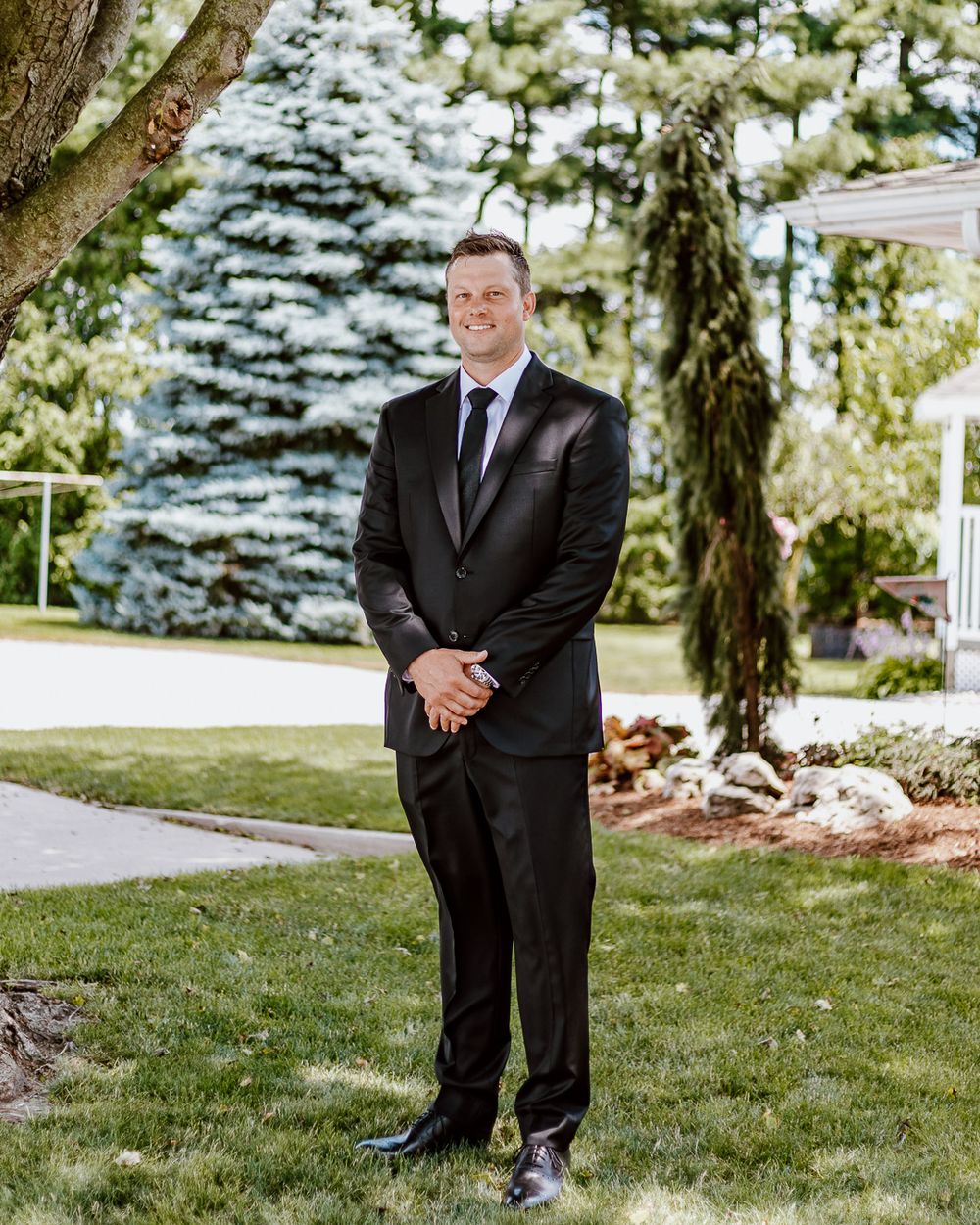 Groom in black suit standing on lawn with blue spruce tree in background.