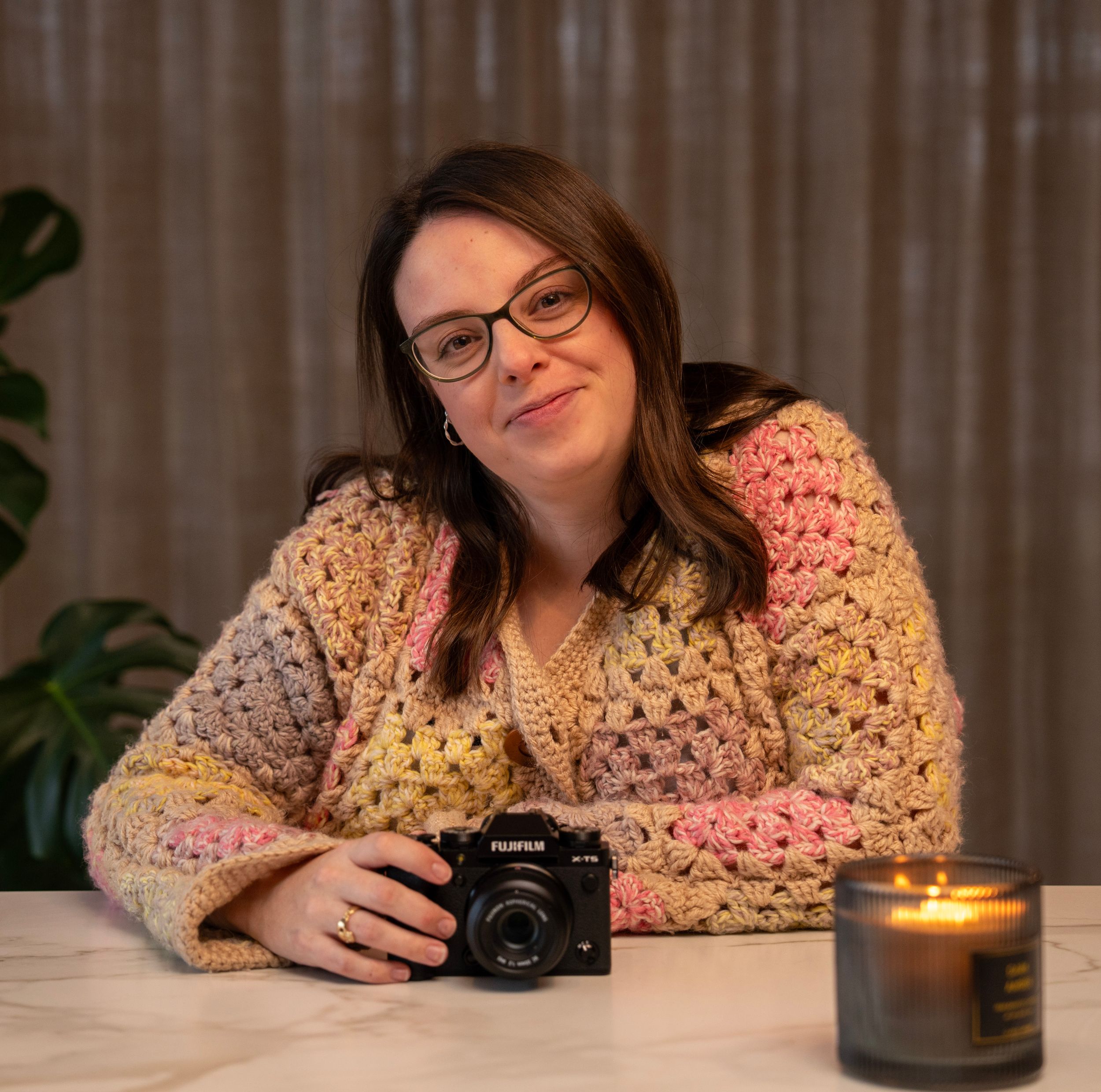 Person in floral top rests at table with camera and lit candle creating cozy indoor atmosphere.