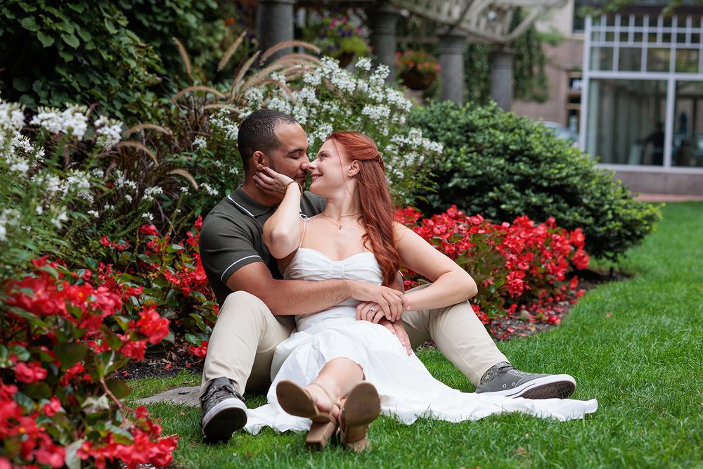 A couple in wedding attire shares romantic moments in a garden setting with blooming red flowers and greenery.