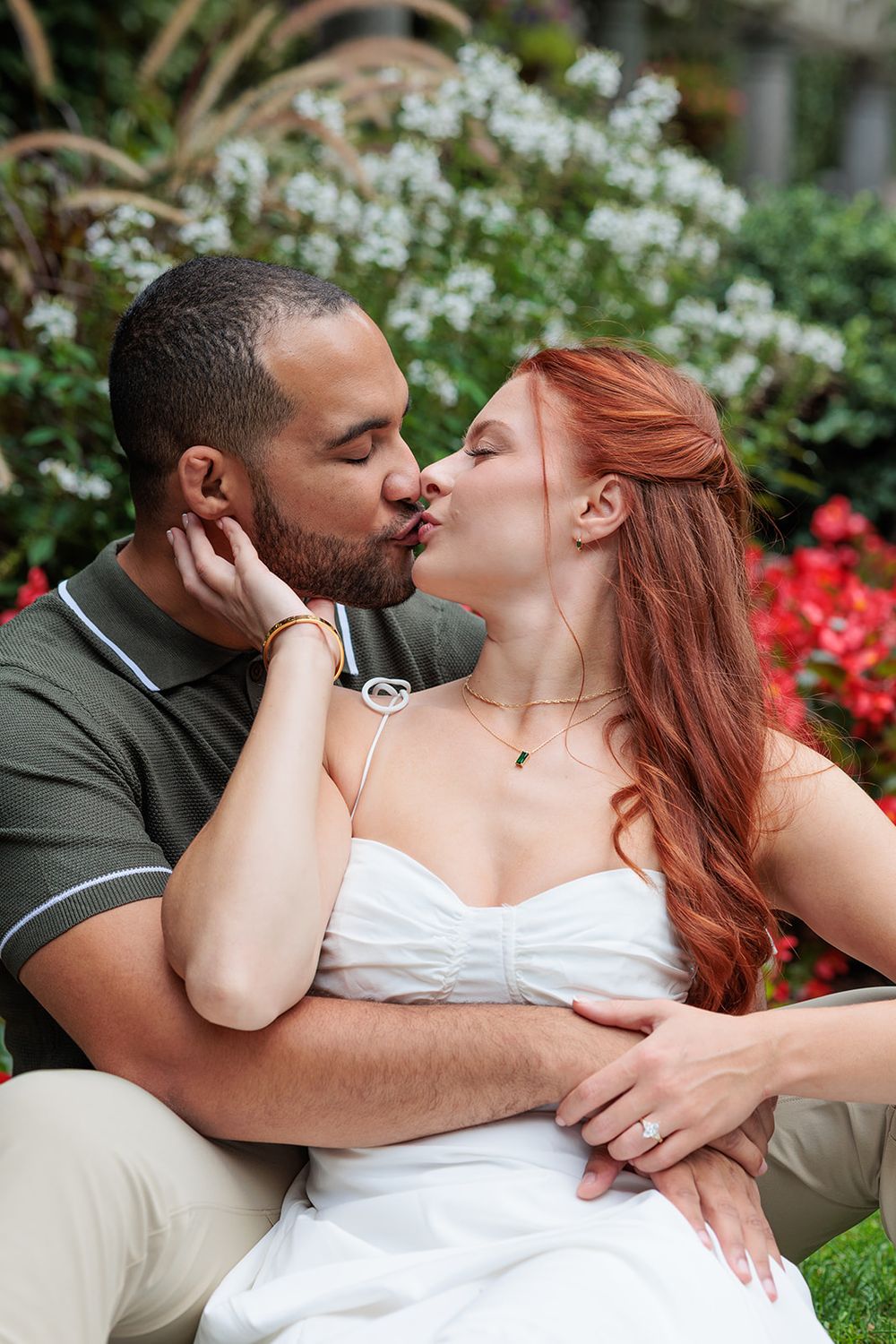 A couple shares intimate kisses in a garden setting with blooming white and red flowers in the background.