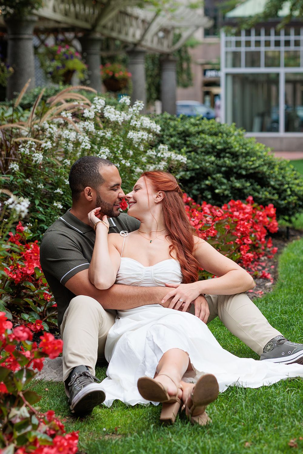 A couple shares an intimate moment while sitting on grass surrounded by blooming red and white flowers in a garden setting.