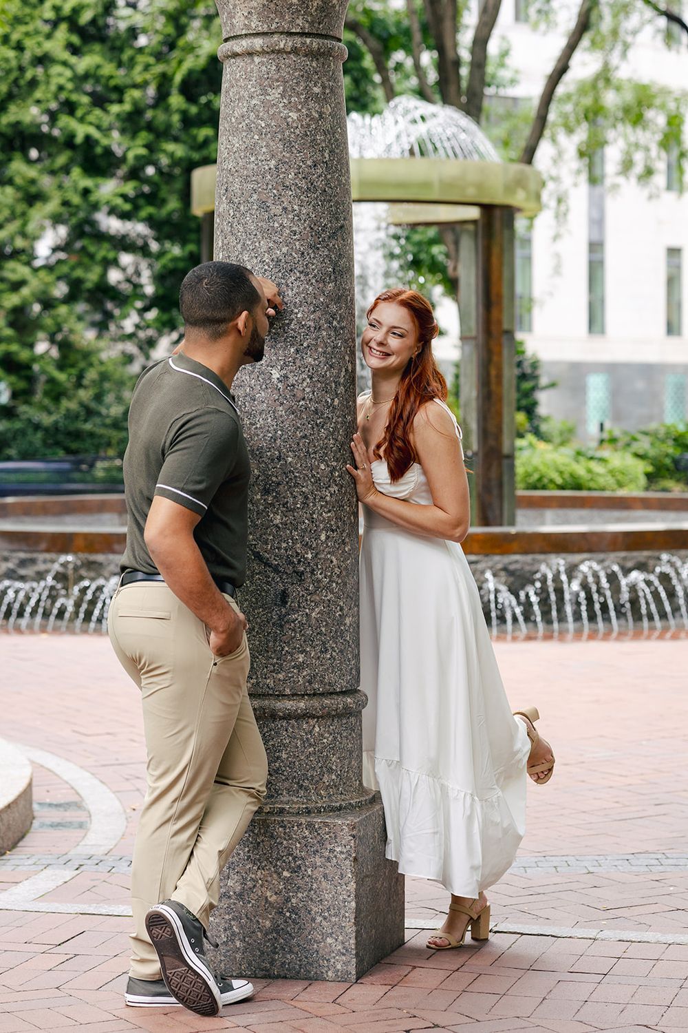 A couple shares a playful moment around a stone column in a courtyard with fountains and greenery.