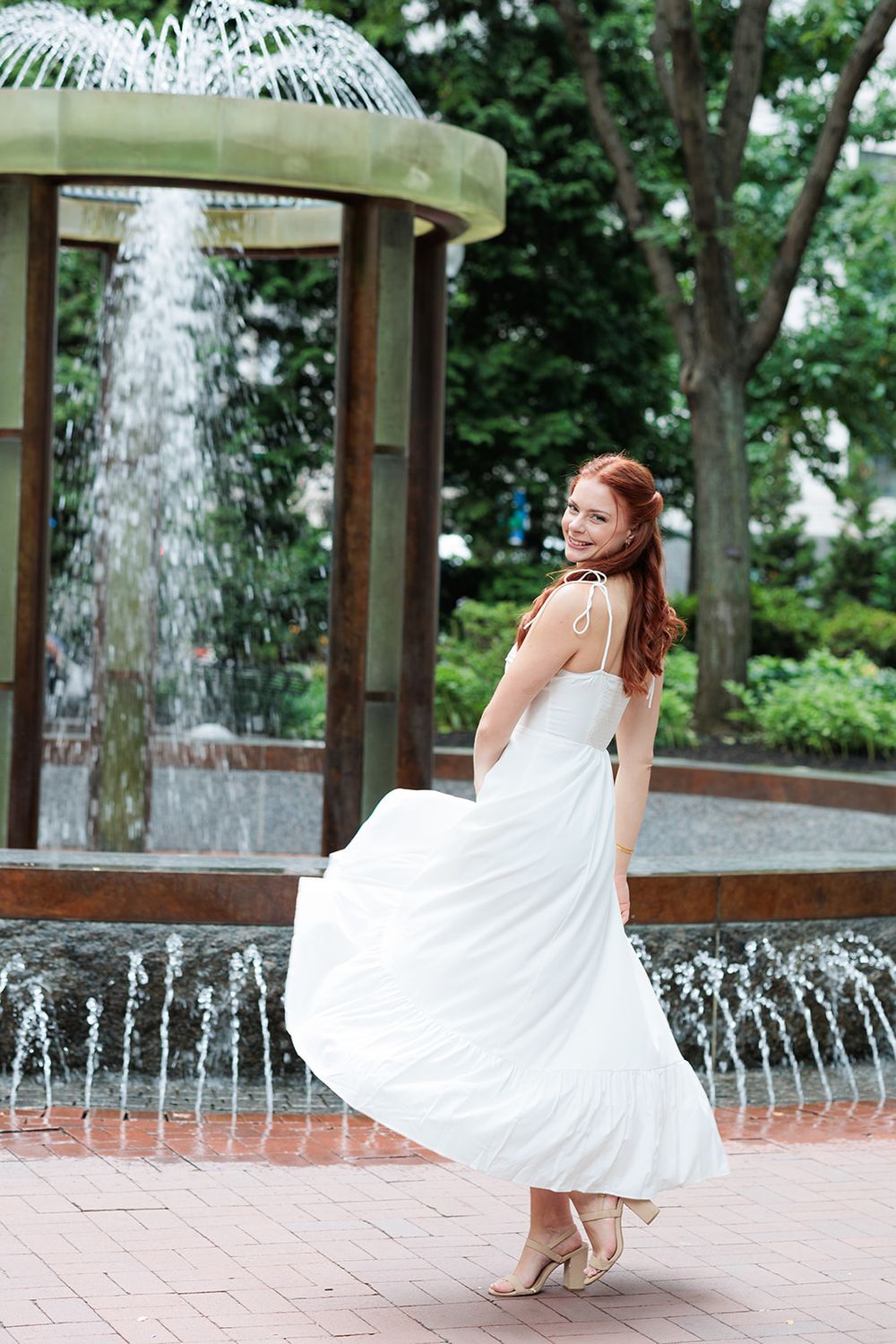 Woman in flowing white dress twirls gracefully by an outdoor fountain on brick pavement.