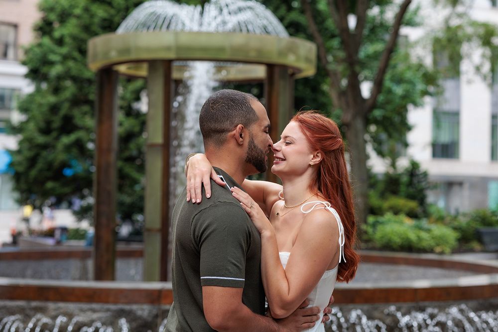 A couple shares a romantic kiss in a park setting with trees and buildings in the background.