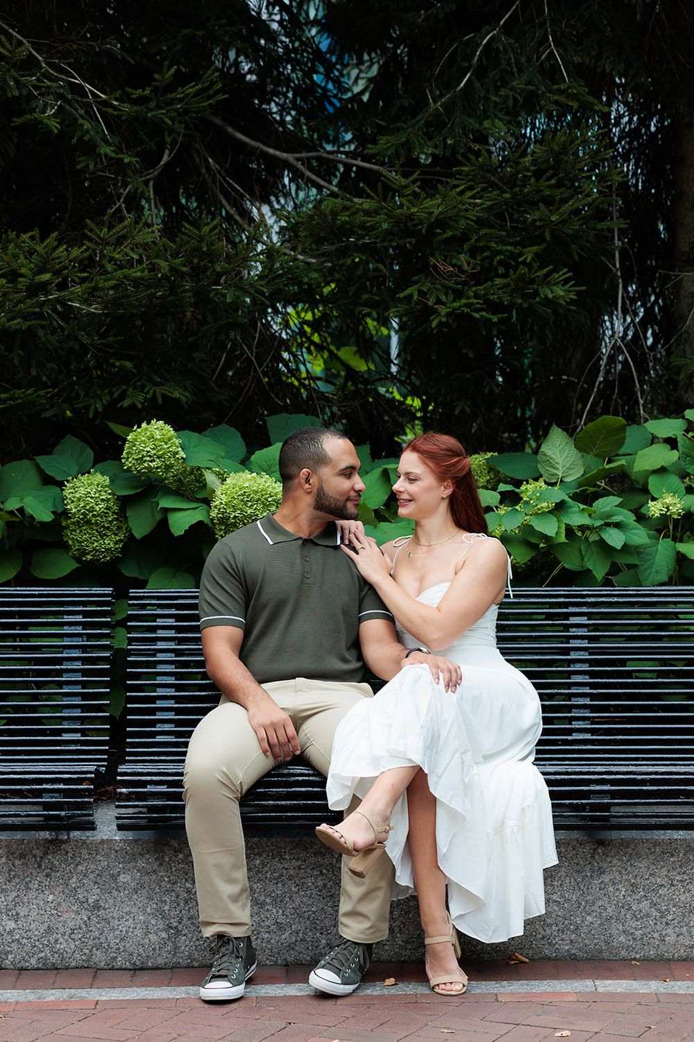 A couple sits together on a bench surrounded by lush greenery and hydrangea blooms in a romantic outdoor setting.
