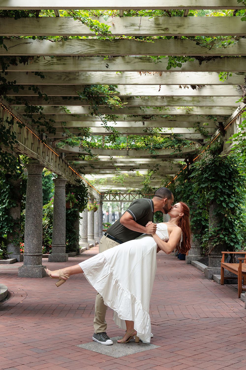 A romantic couple shares a dip kiss moment under a vine-covered pergola on a brick patio.