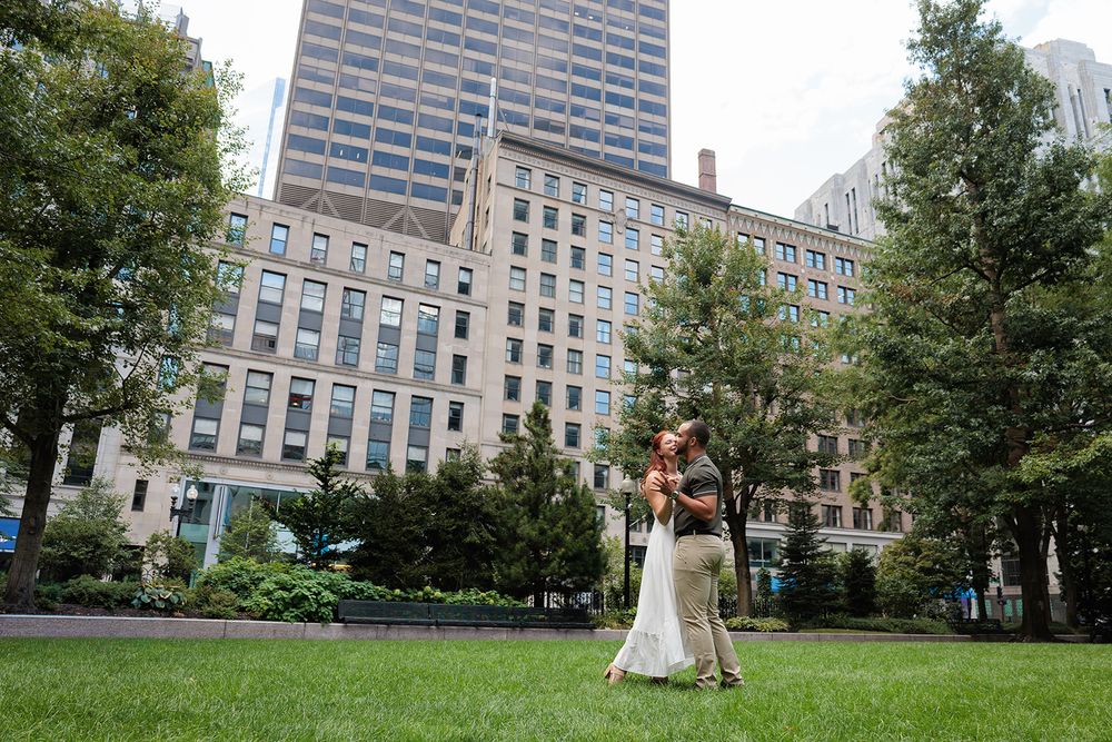 A couple shares a romantic moment on a grassy lawn surrounded by tall city buildings and trees.