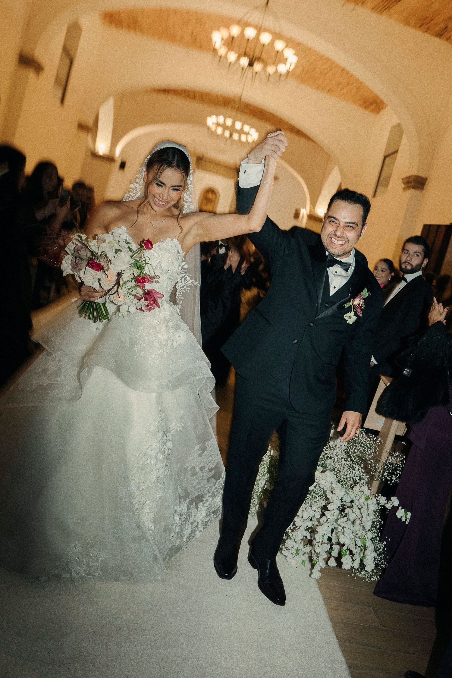 A joyful wedding couple celebrates during their reception, with the bride in a layered white gown and groom in classic tuxedo.