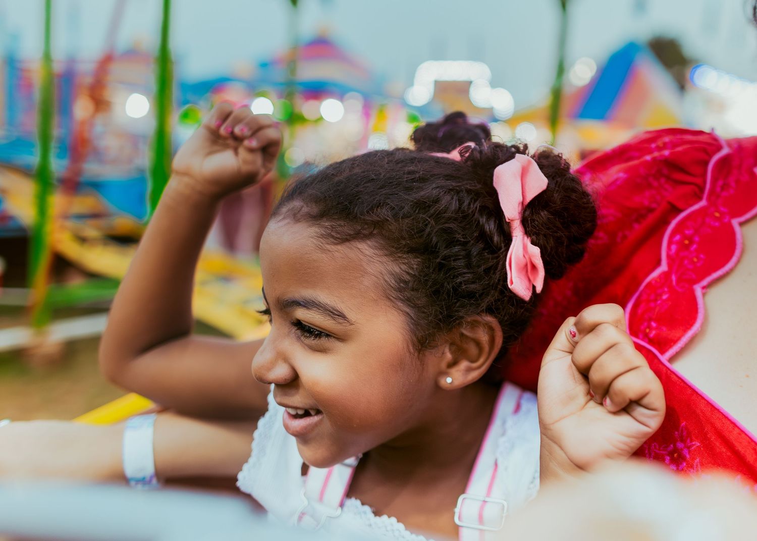 Child in a pink bow laughing and playing at a colorful playground with their arms raised in joy.