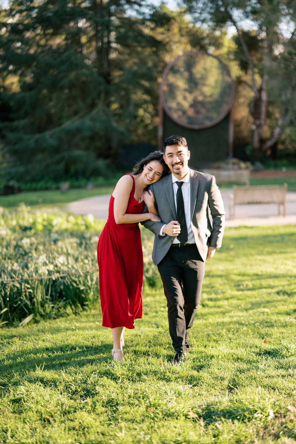 A couple in formal attire playfully walks through a sunlit garden with green grass and trees.