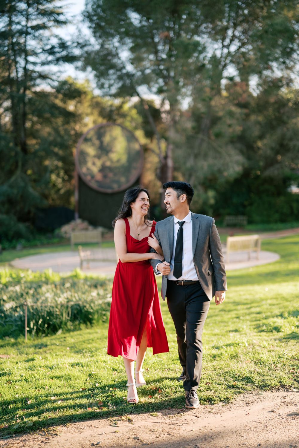 A couple walks together on a sunlit dirt path through a green park, the woman wearing a flowing red dress.
