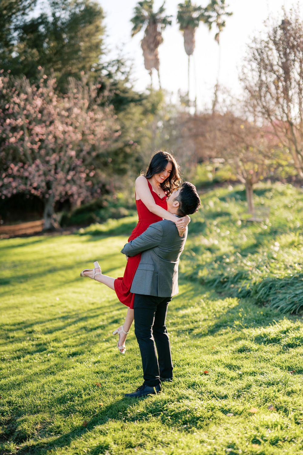 A romantic moment captured in a sunlit garden with blooming cherry blossoms and palm trees in the background.