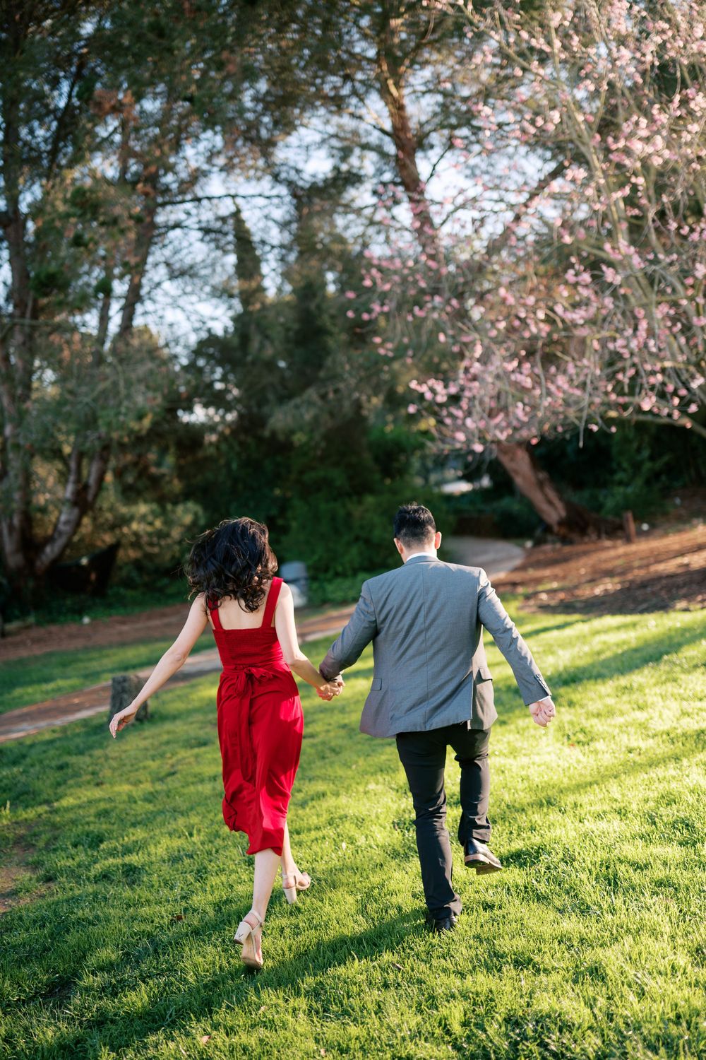 A couple runs playfully through a grassy field under blooming cherry blossom trees on a sunny afternoon.