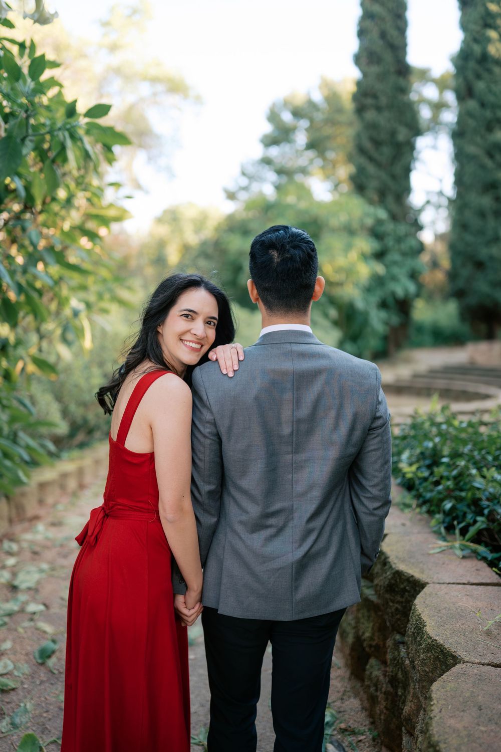 A couple in a red dress and grey suit share a romantic moment on a garden path surrounded by greenery.
