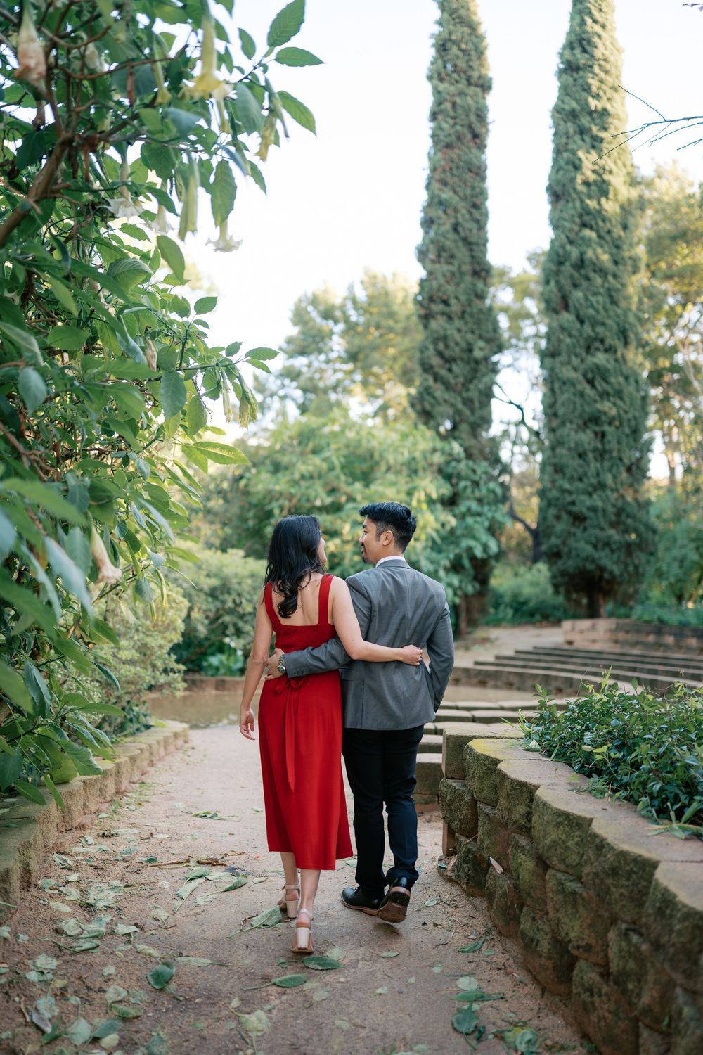A couple walks together on a garden path with tall cypress trees and lush greenery in the background.