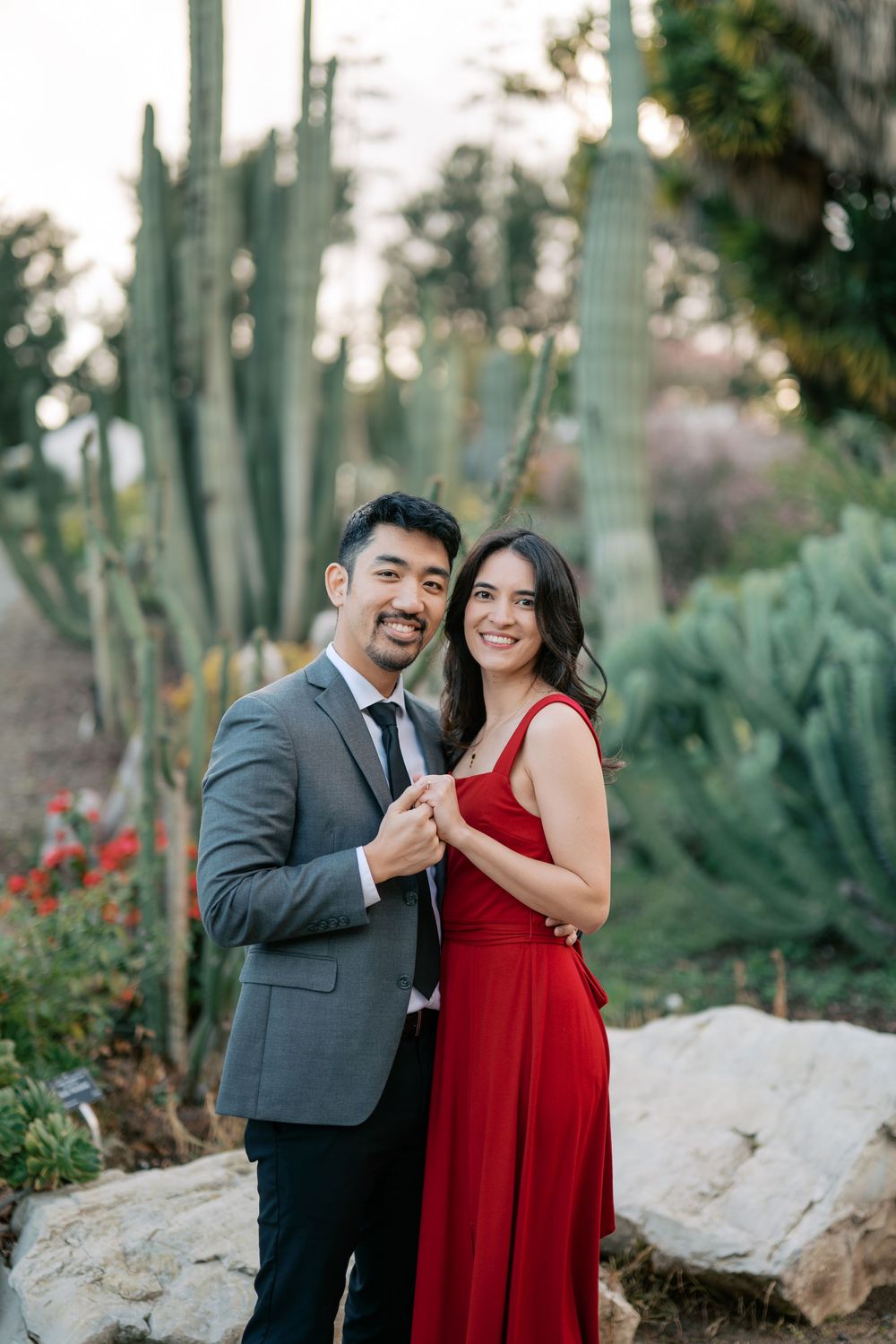 A romantic outdoor photoshoot featuring a couple in a red dress and grey suit among desert plants.
