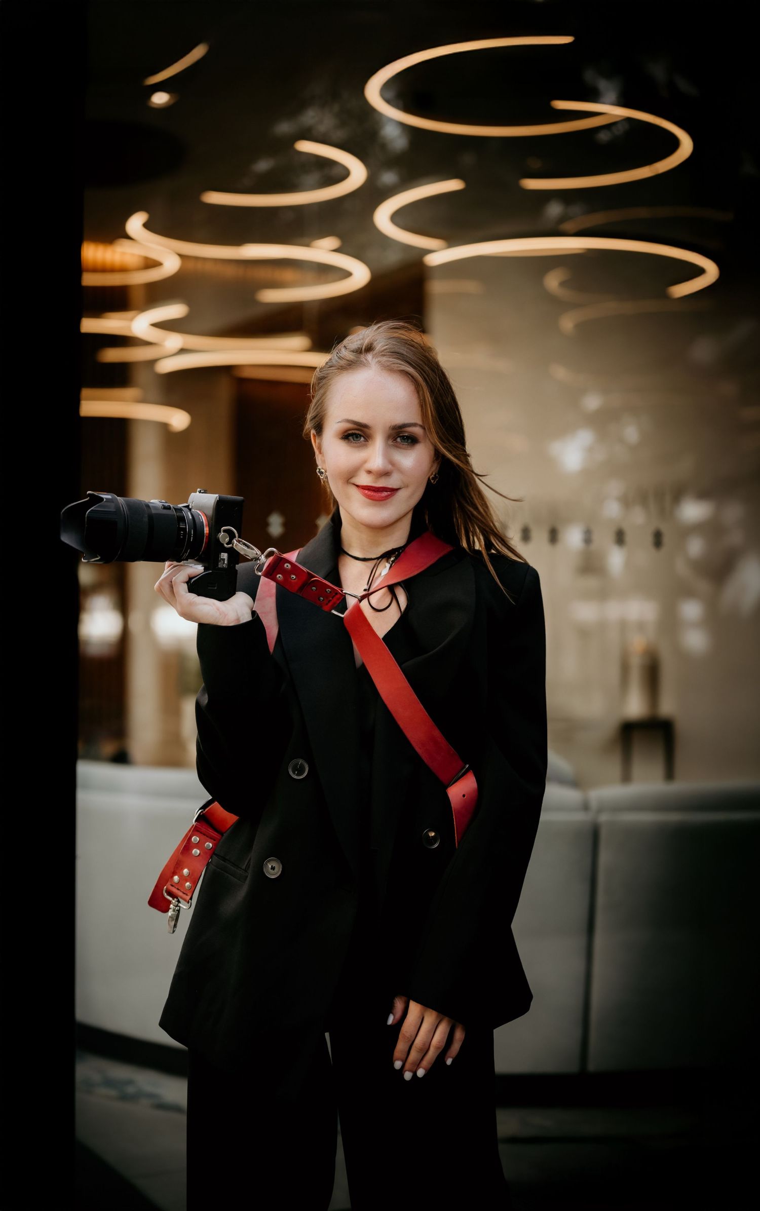 Photographer in black coat with red camera strap stands beneath swirling light trails in moody indoor setting.