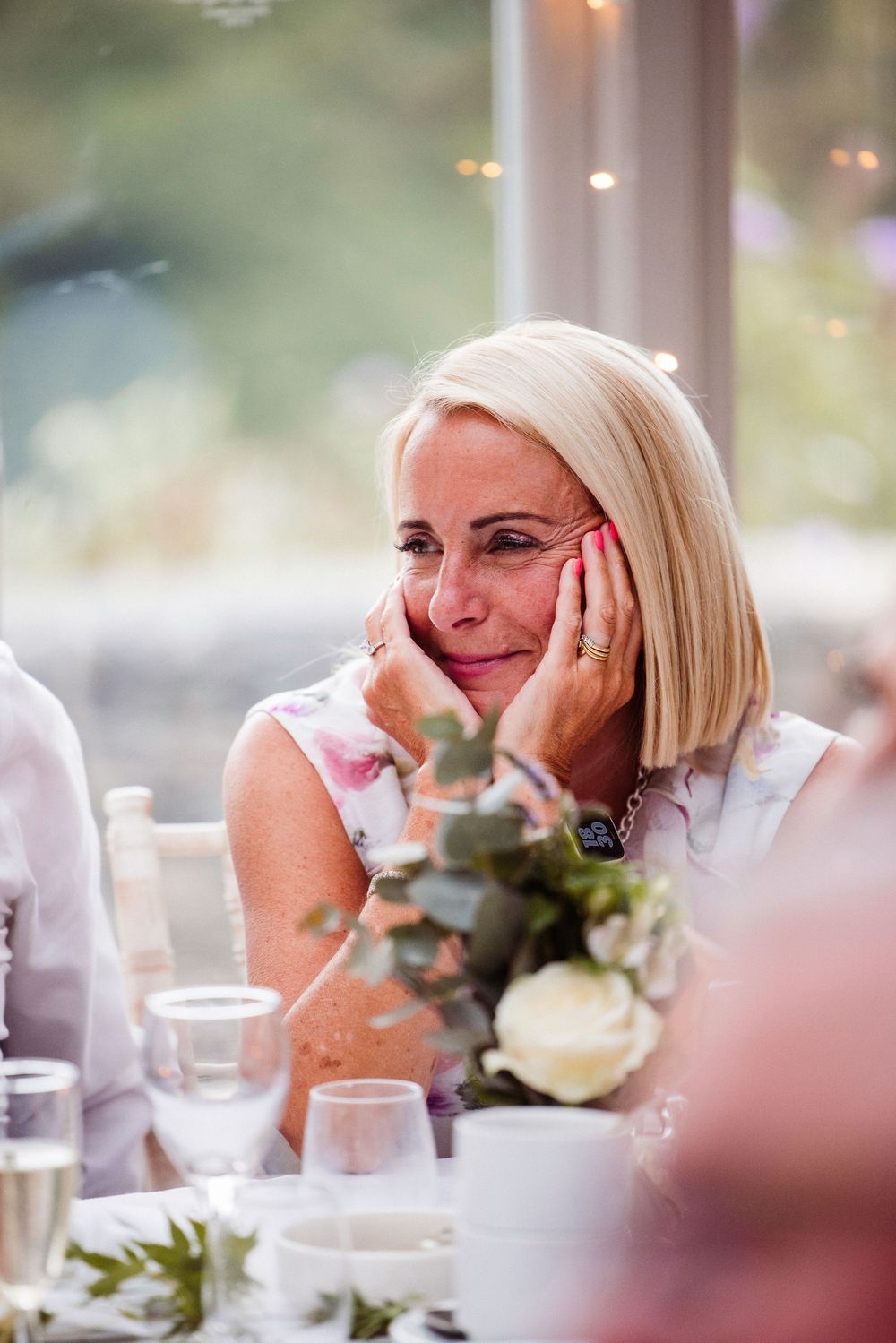 A wedding guest in a white dress rests her head in her hands while seated at a reception table with floral centerpieces.