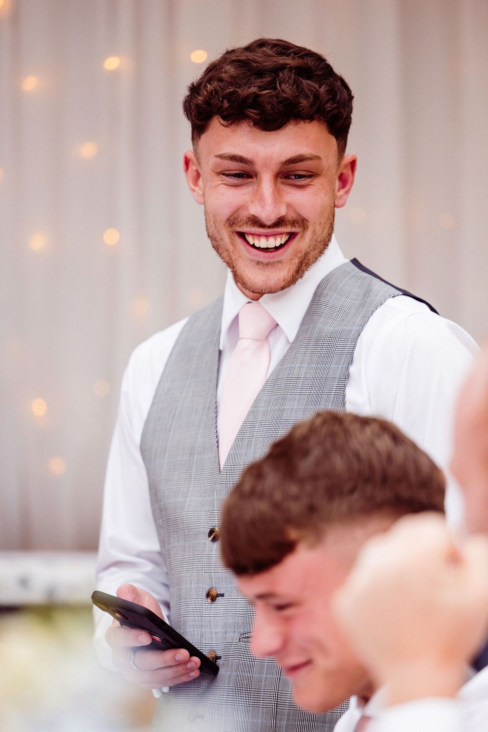 A person in a light gray vest and pink tie smiles while giving a speech at a formal event.