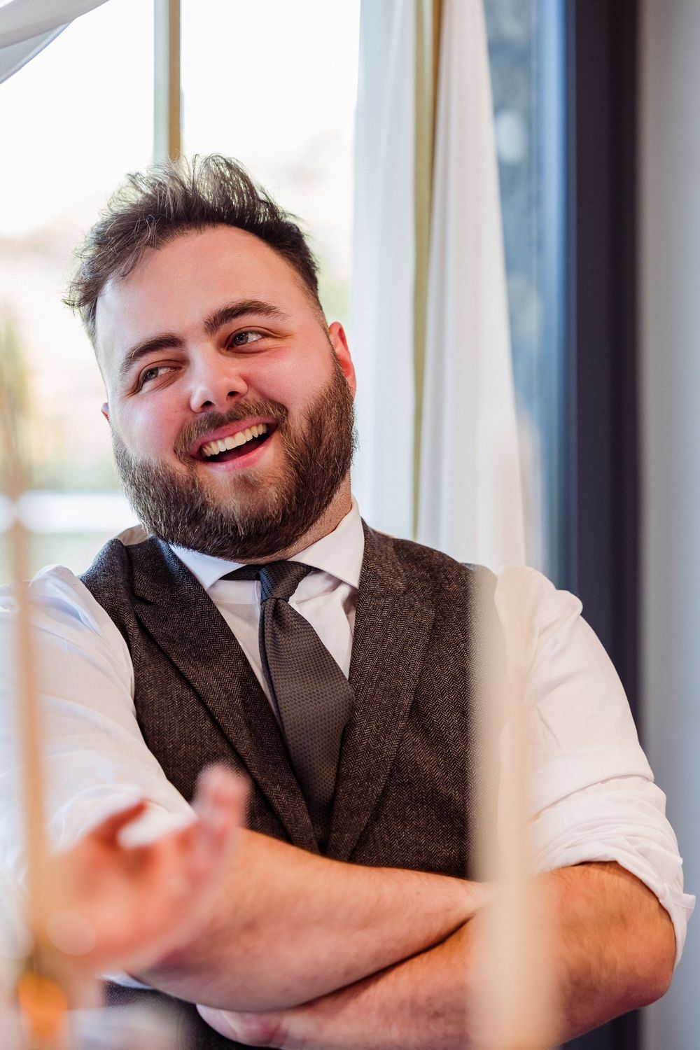 A person in formal attire with a brown vest and tie smiles warmly while sitting with arms crossed.