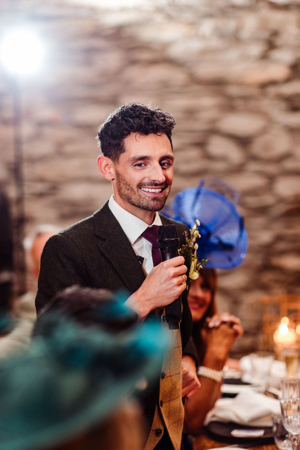 A person in a black suit gives a speech while holding a microphone at an elegant candlelit dinner event.