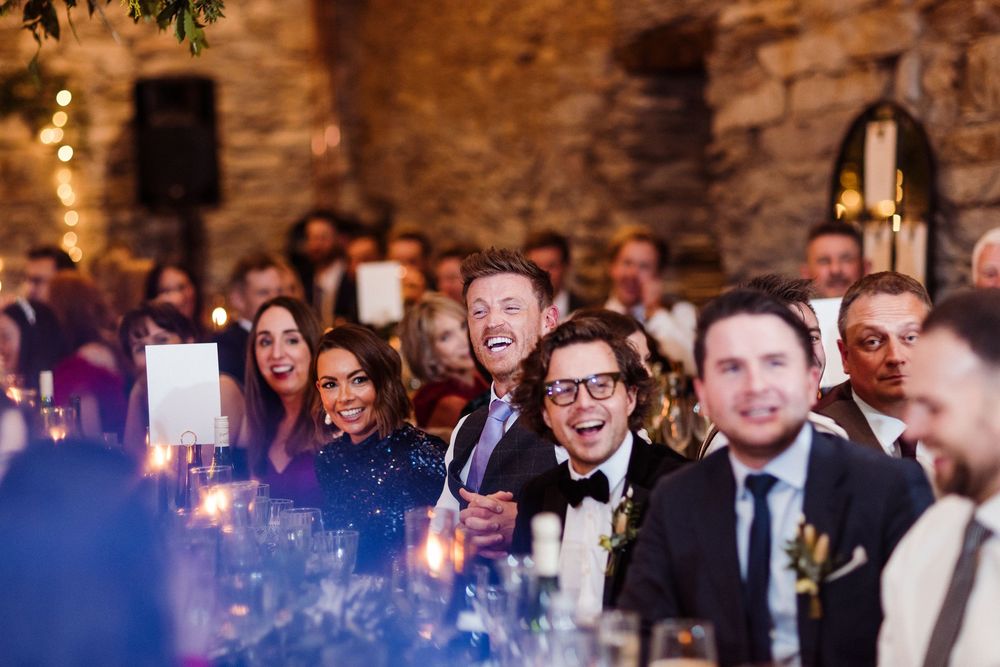 Wedding guests laugh and react during reception speeches at rustic stone venue with candlelit ambiance.