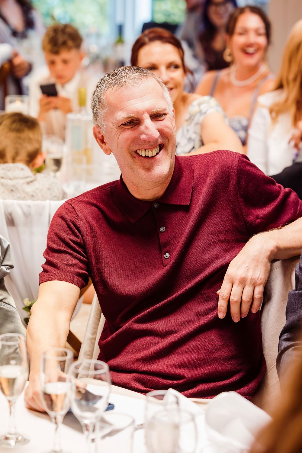 A person in a burgundy polo shirt smiles while seated at a formal dining table during an event.