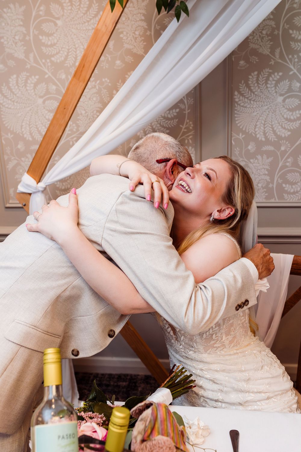 Joyful couple embraces and laughs during wedding reception with wine bottles and table decor visible in foreground.