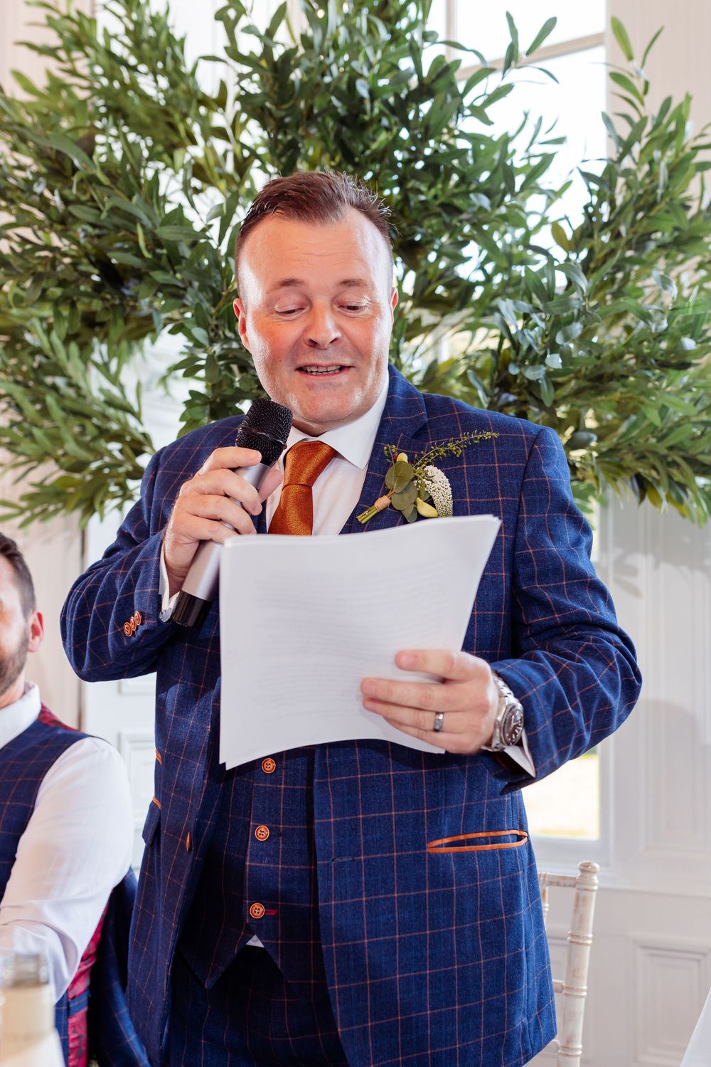 A speaker in a navy plaid suit and orange tie reads from papers during a formal event with greenery in the background.