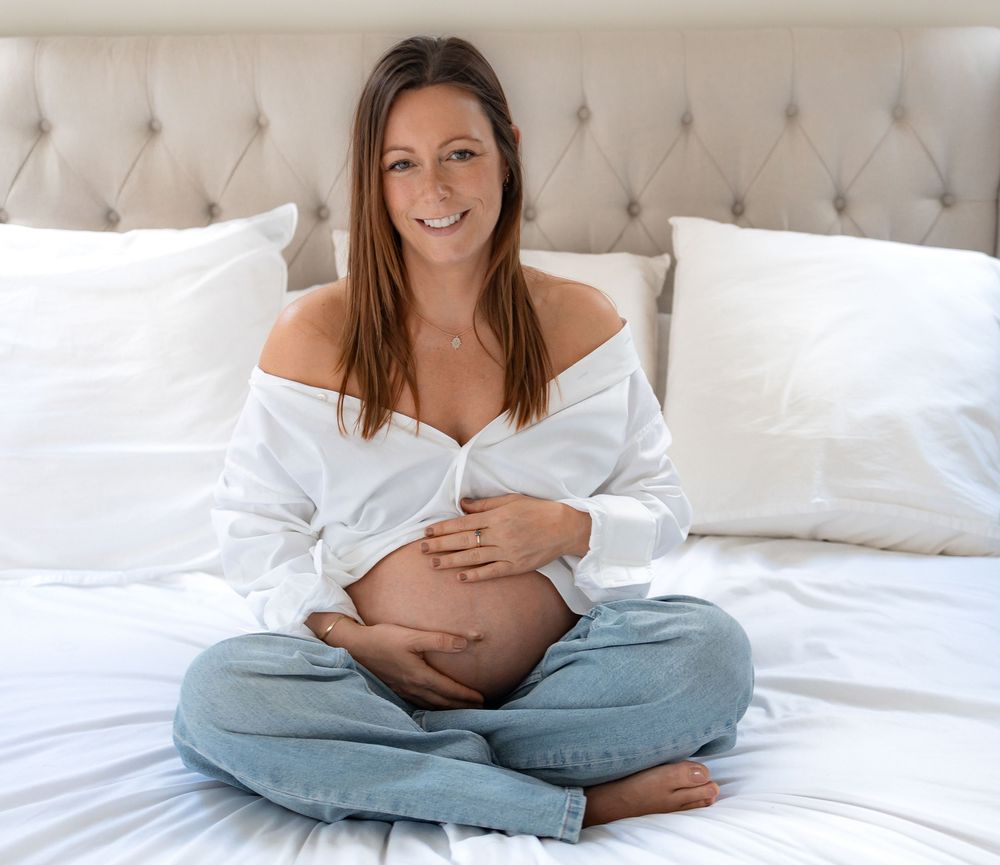 Smiling pregnant woman sitting cross-legged on white bed, wearing open white shirt and jeans, cradling baby bump.