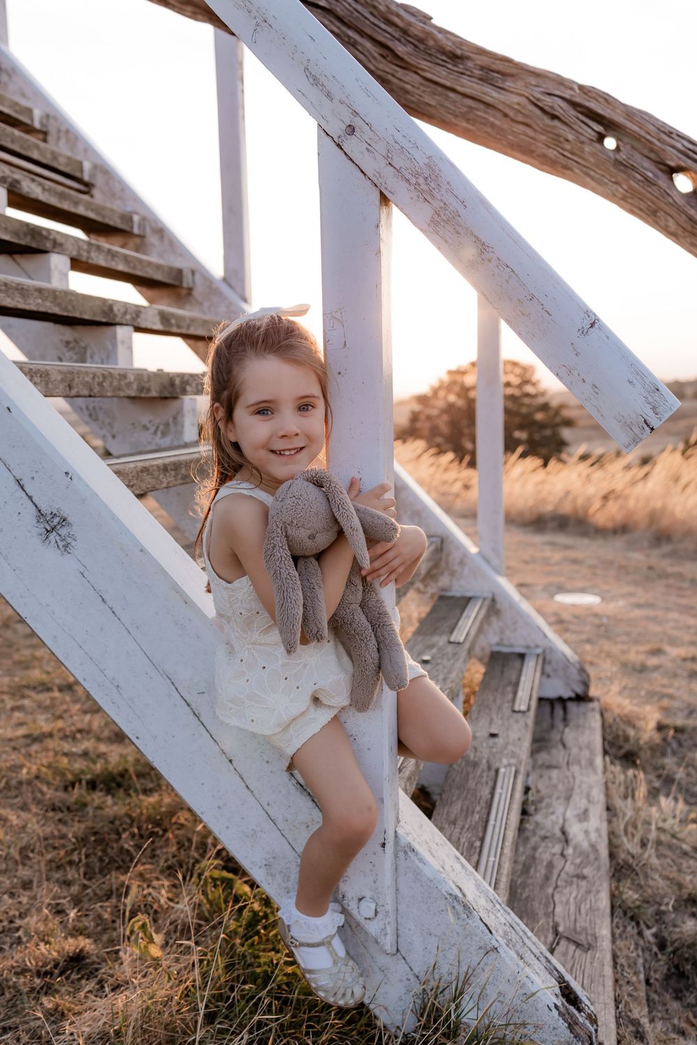 Young girl holding stuffed bunny toy on white wooden stairs at golden sunset in rural field.
