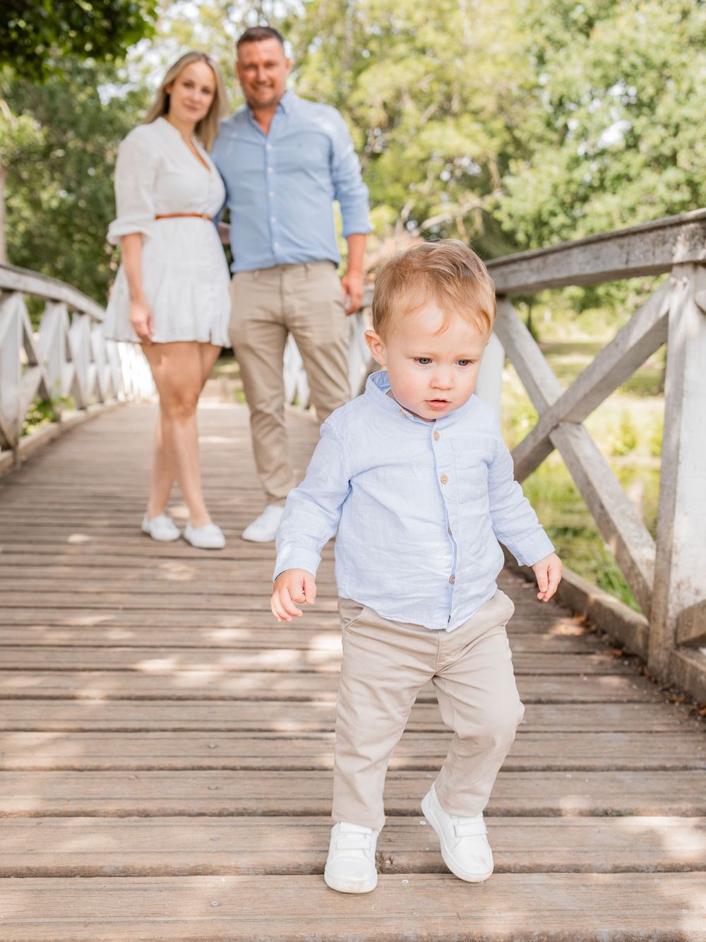 A toddler boy in a light blue shirt walks on a wooden bridge while his parents hold hands and smile behind him.