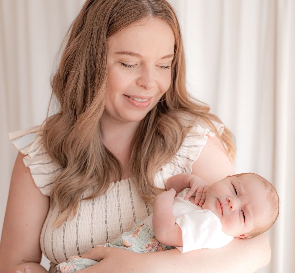 A smiling young mother holds her sleeping newborn baby in her arms in a soft, bright studio setting.