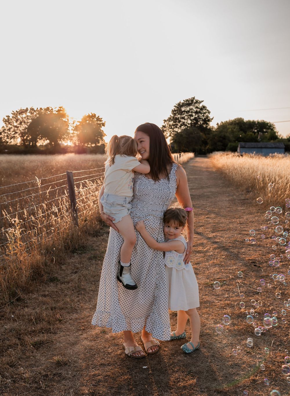 A mother holds a toddler while another child hugs her in a golden sunlit field at sunset.