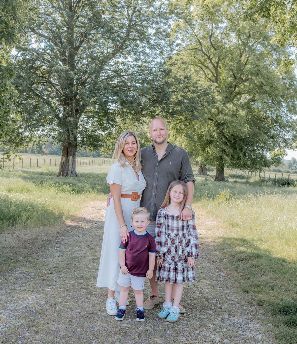 Family of four posing on a gravel path surrounded by lush green trees and meadows on a sunny day.