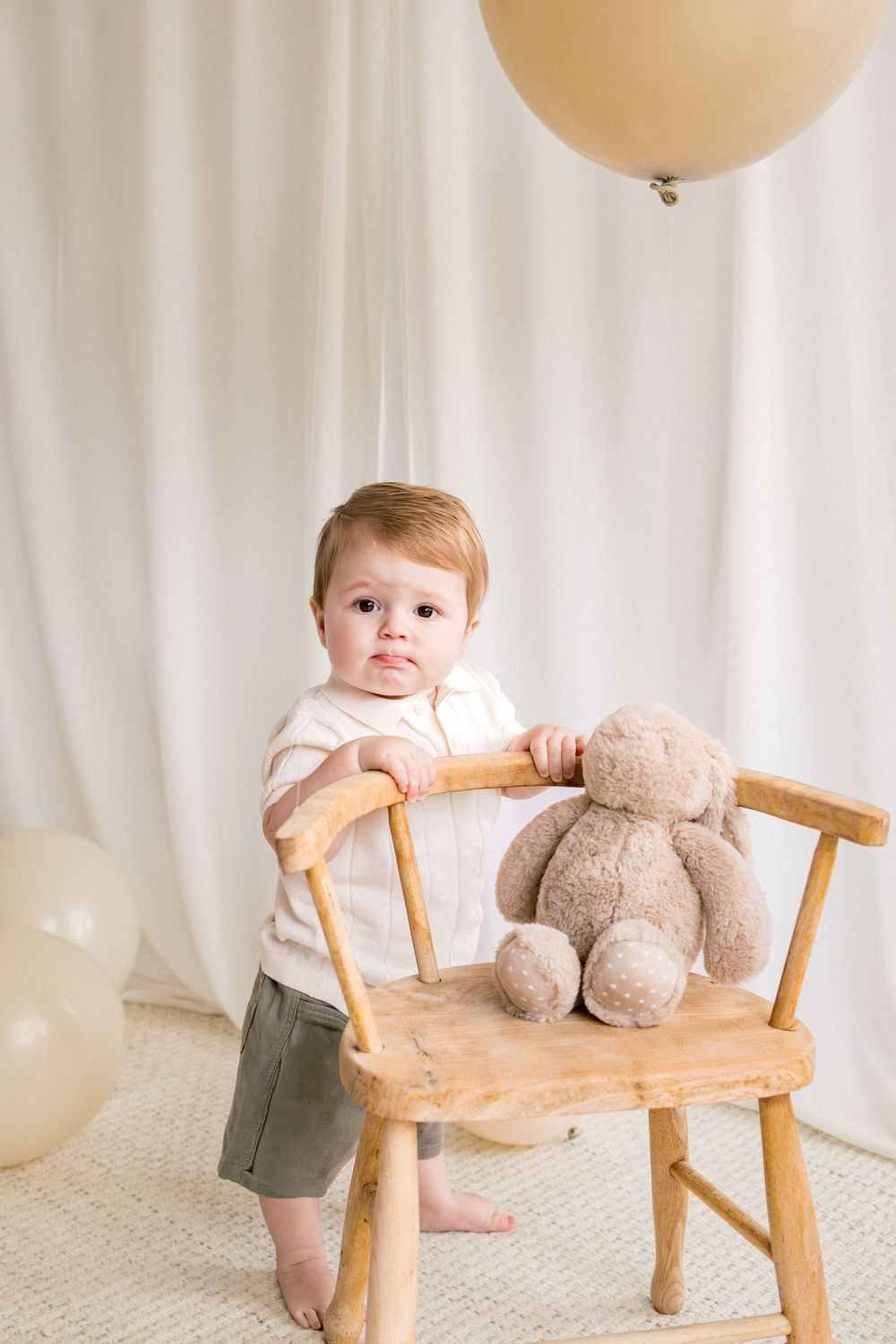 Toddler boy standing beside wooden chair with teddy bear, surrounded by neutral balloons on white backdrop.