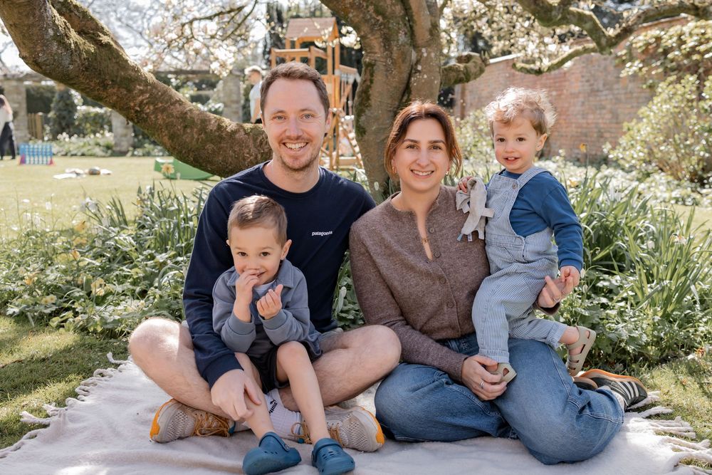 Happy family of four sitting on a blanket outdoors under a large tree in a blooming garden on a sunny day. Photography in Buckinghamshire.